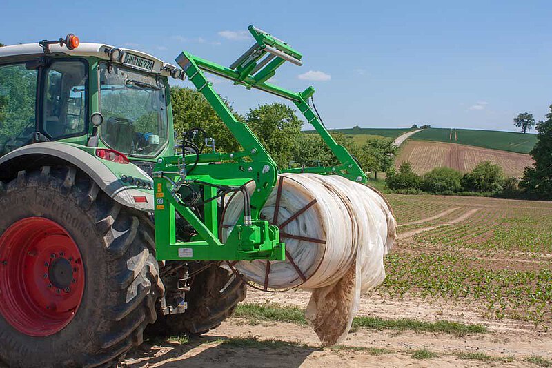 A green tractor with a yellow trailer on farmland under a bright blue sky with clouds.